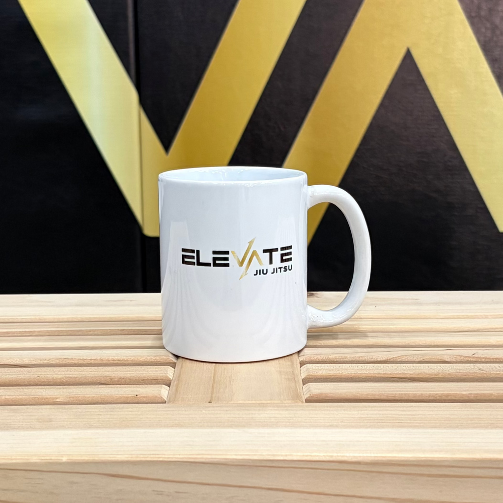 White mug with 'Elevate' branding on a wooden surface in front of a black wall with 'ELEVATE' letters.