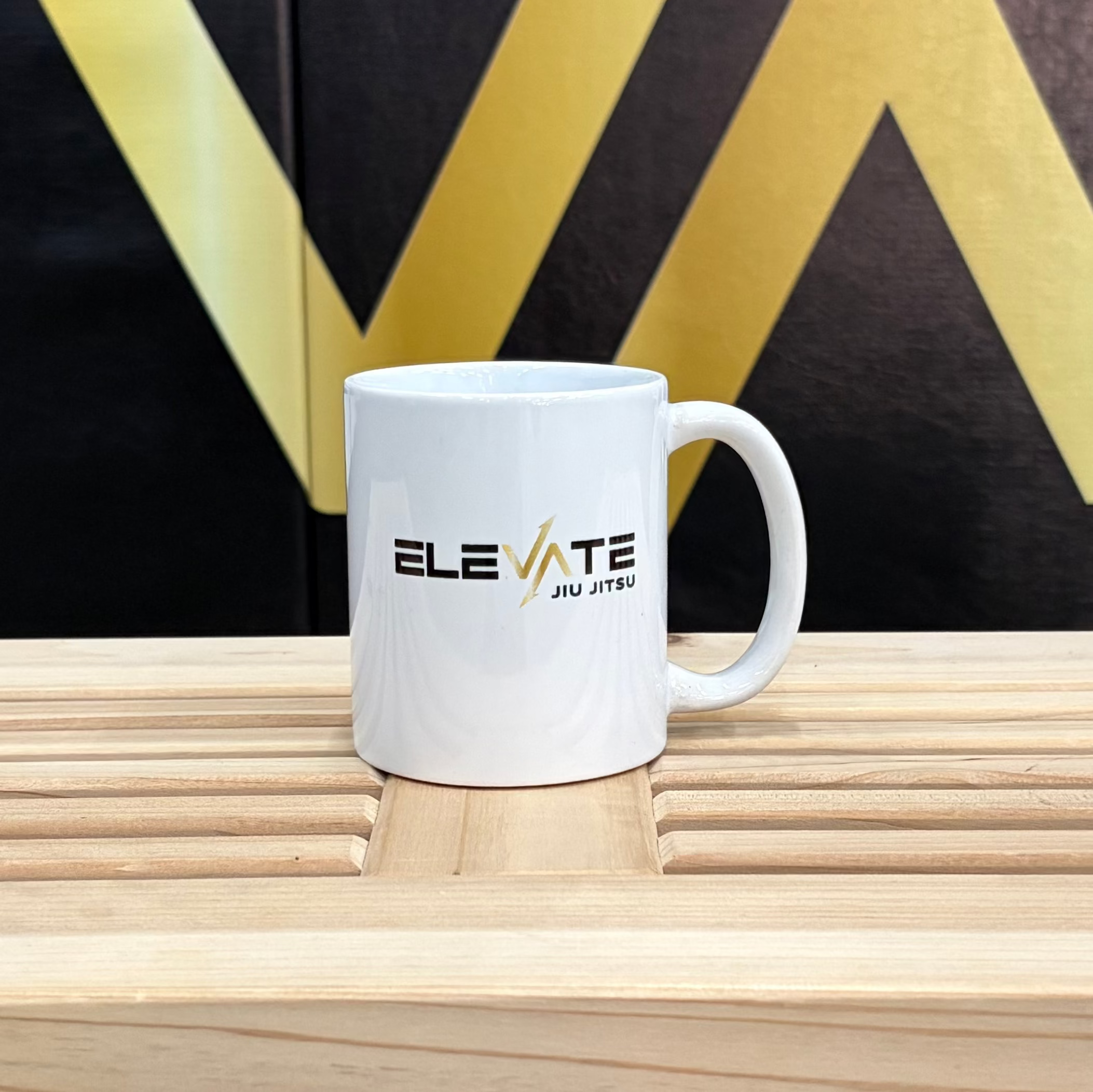 White mug with 'Elevate' branding on a wooden surface in front of a black wall with 'ELEVATE' letters.