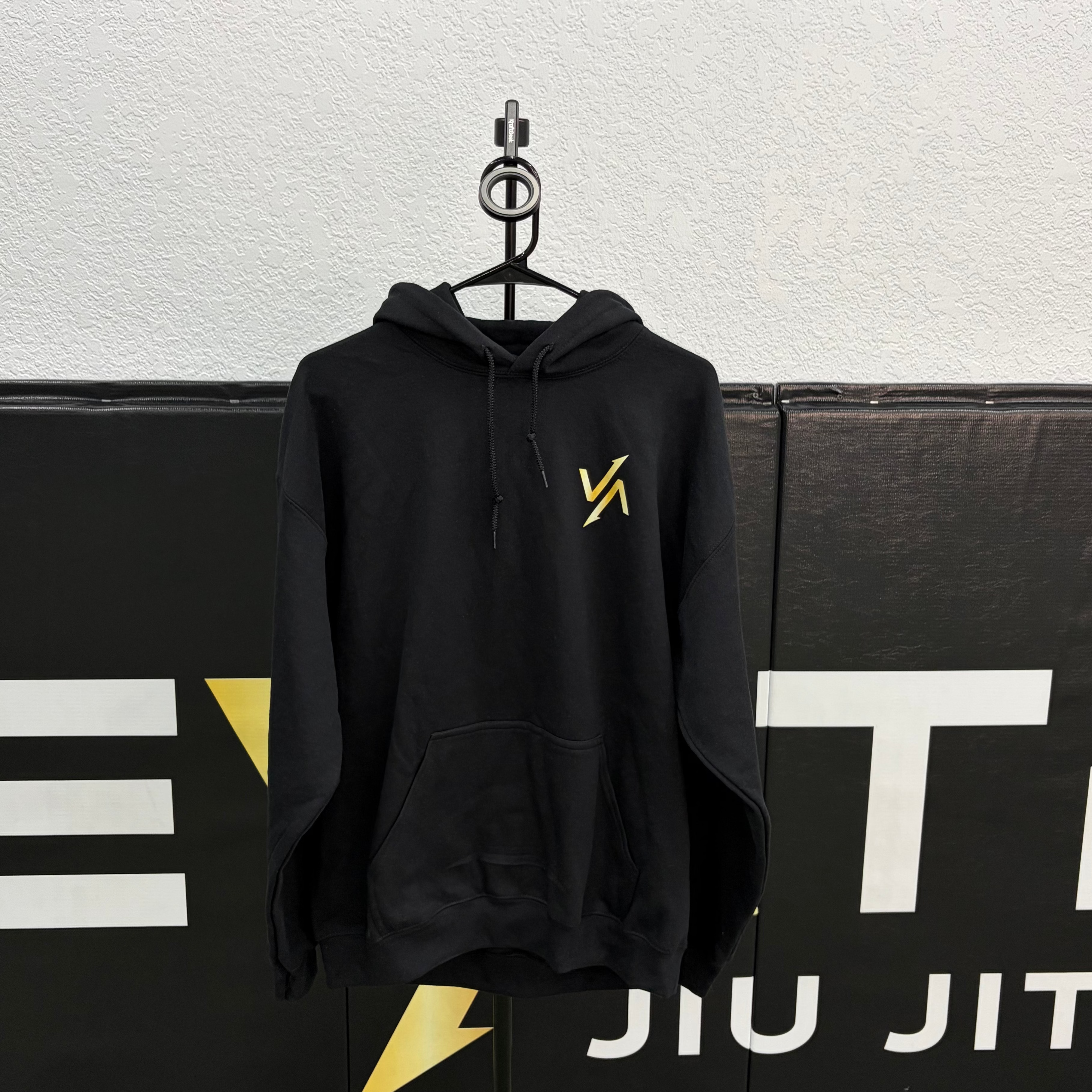 Black hoodie with a yellow logo on a hanger against a Brazilian and American flag background.